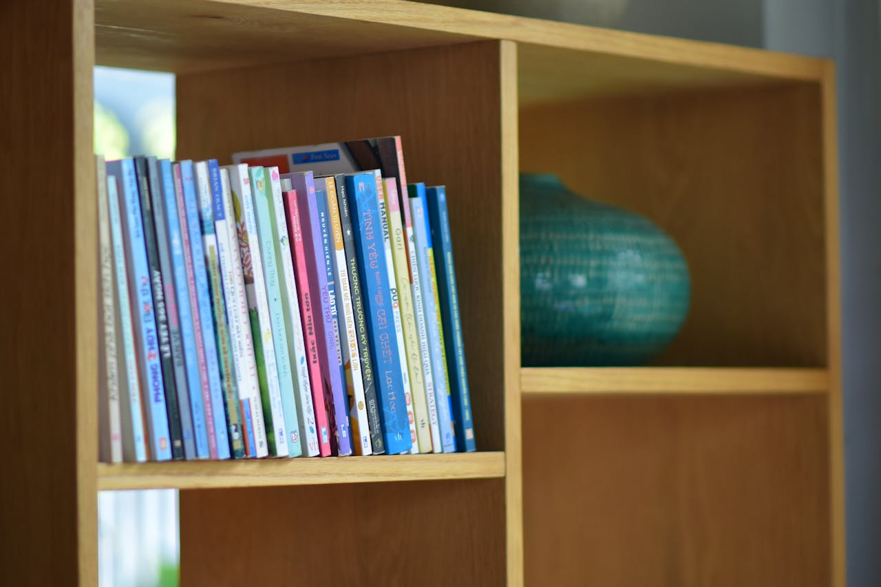 A collection of colorful childrens books arranged on a wooden bookshelf with a decorative vase.